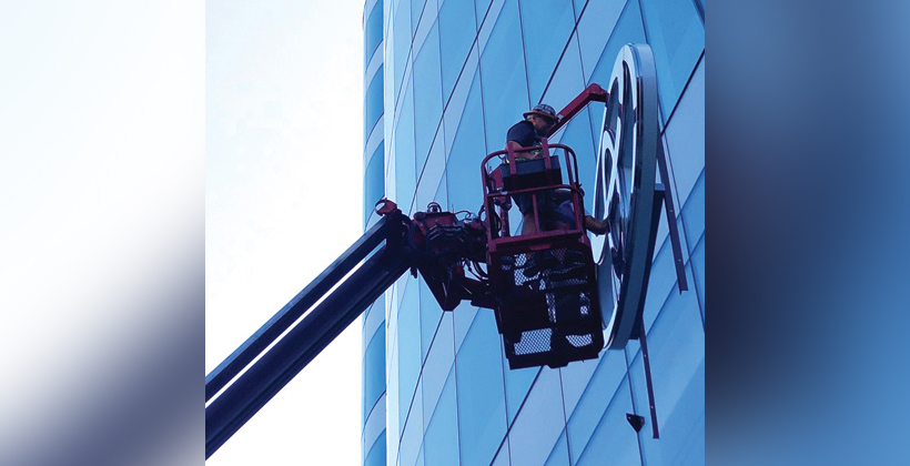 An installer assembles a sign while standing on a crane.
