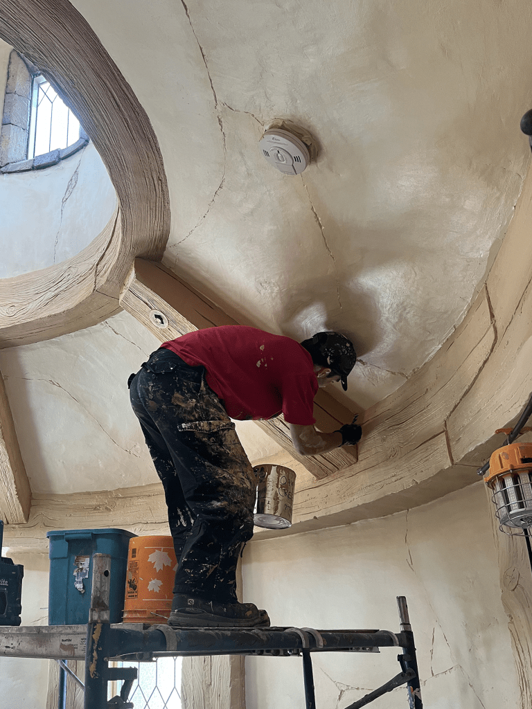 Worker hand-finishing a curved plaster ceiling inside a fantasy-themed room under construction, standing on scaffolding