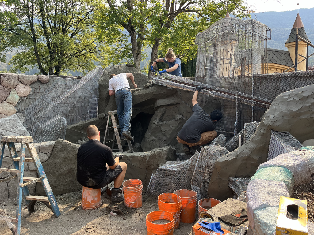Four workers applying concrete over wire mesh to sculpt artificial rock formations outdoors, with a castle turret visible in the background