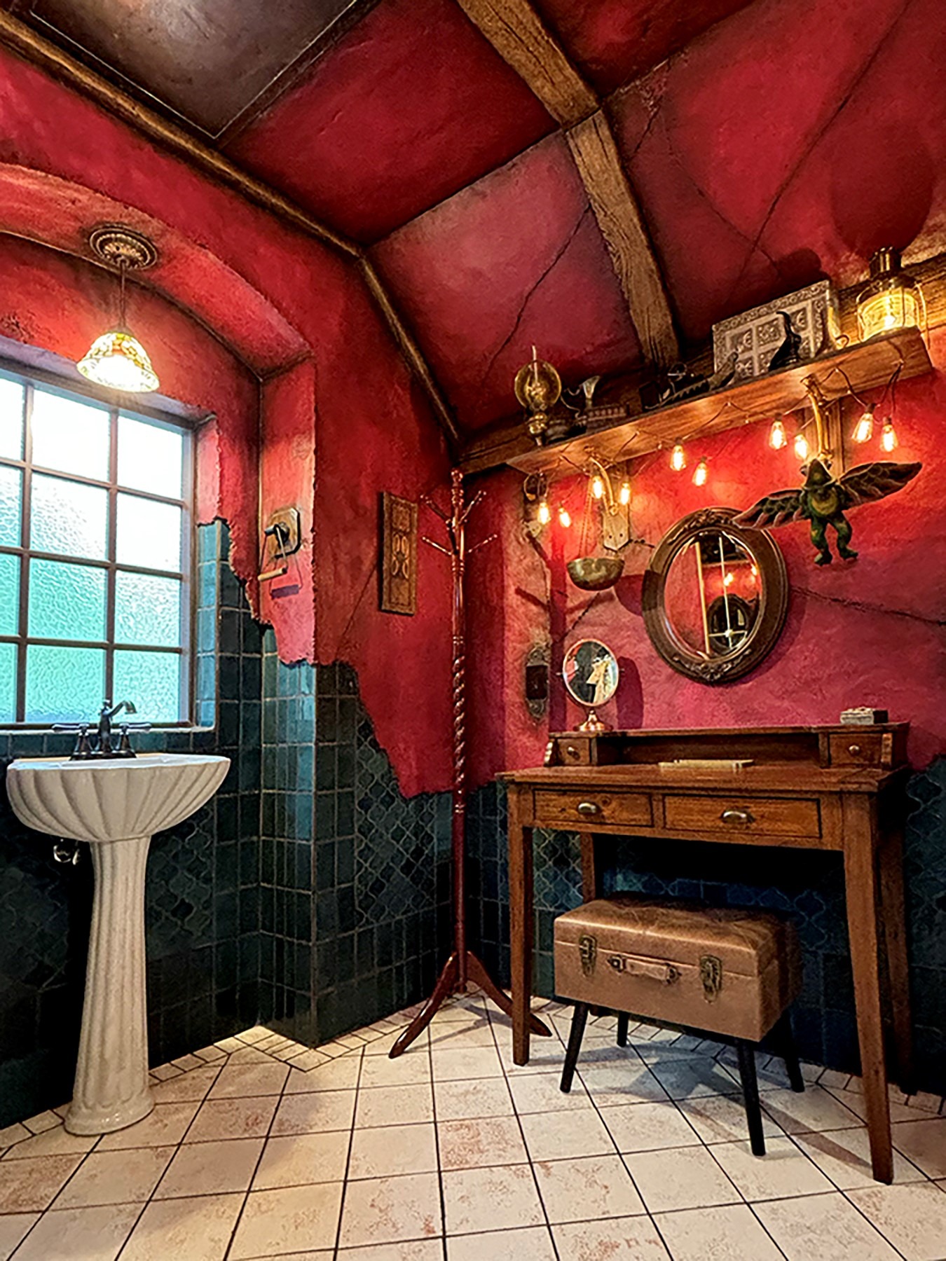 Fantasy-themed bathroom with red textured walls, green tile, pedestal sink, and antique writing desk