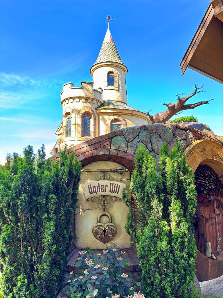 "Under Hill" sign with a heart padlock motif set into a stone arch, with a fantasy castle turret in the background