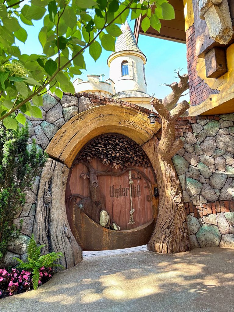 Round hobbit-style wooden door labeled "Under Hill" framed by a carved tree trunk archway and stone wall