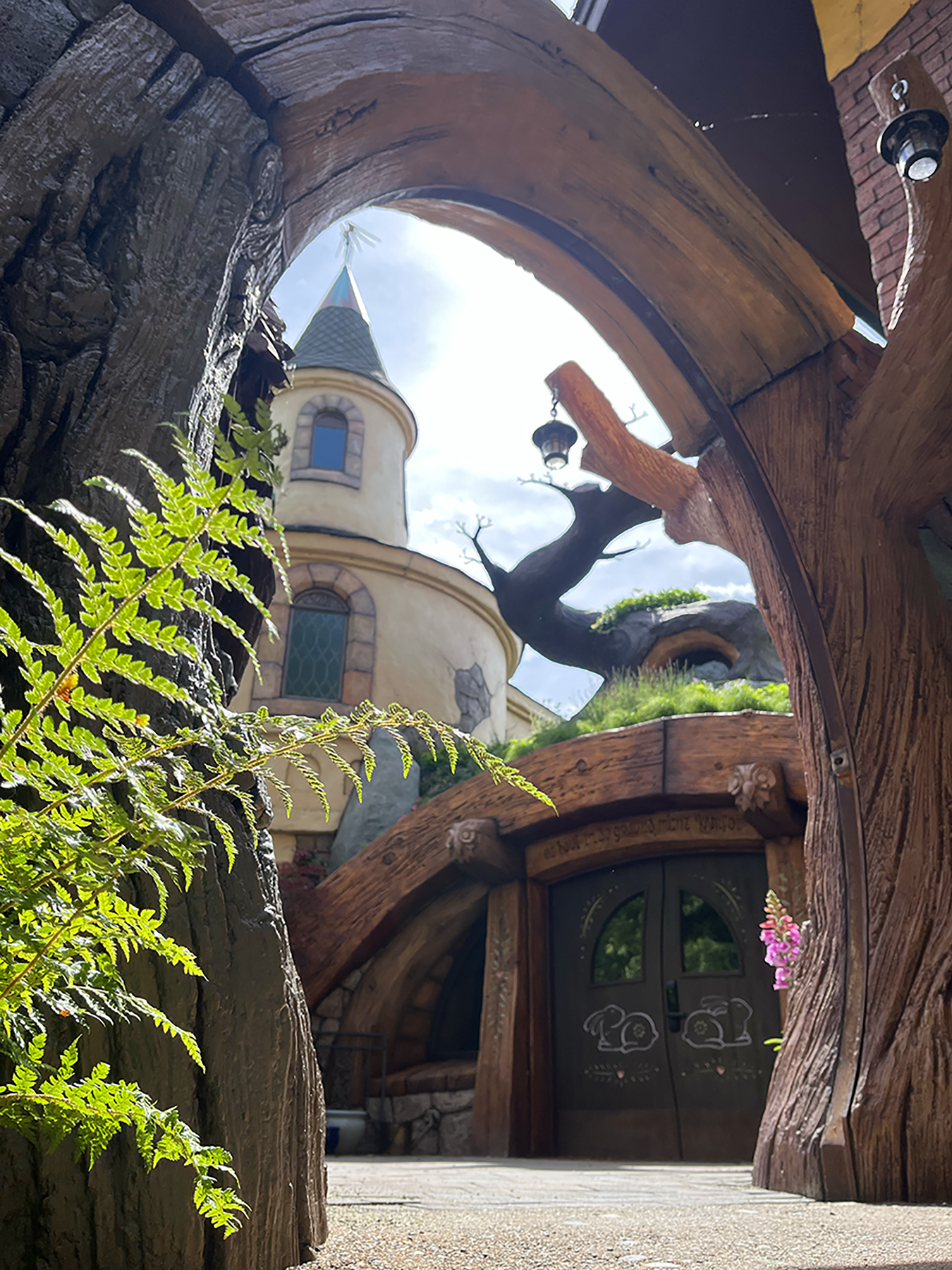 View through a carved wooden tree trunk arch toward a hobbit-style round door and fantasy castle turret beyond