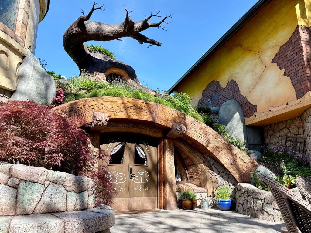 Fantasy hobbit hole entrance with a curved wooden arch door decorated with painted rabbits, a grass-covered roof, and a gnarled tree sculpture above