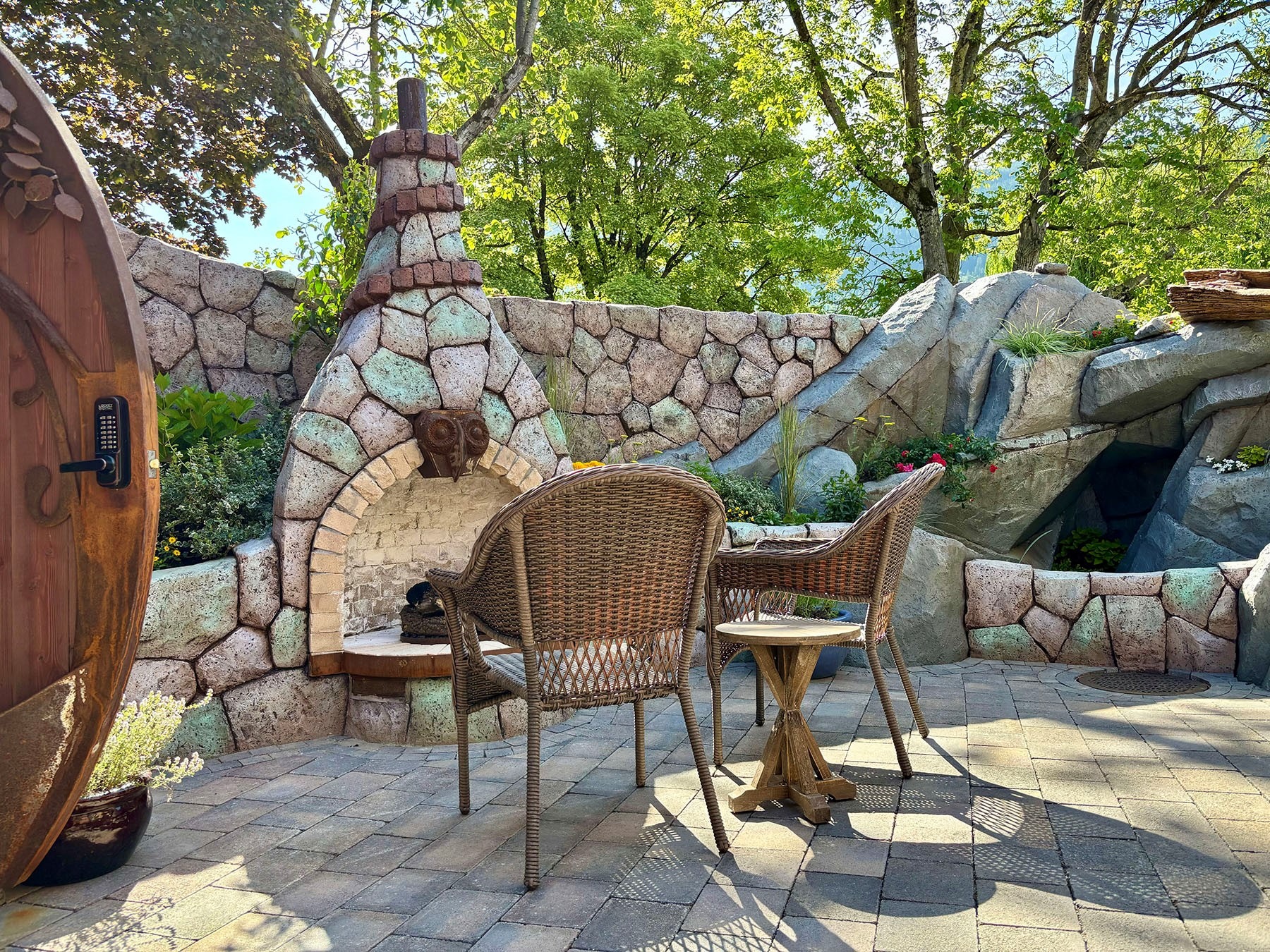 Outdoor patio at a fantasy-themed inn with wicker chairs, a stone fireplace, and boulder-lined garden walls