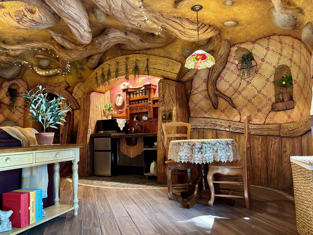 Interior of a hobbit-style room with carved wood-grain walls, floral wallpaper, dining table with lace cloth, kitchenette, and Tiffany pendant lamp