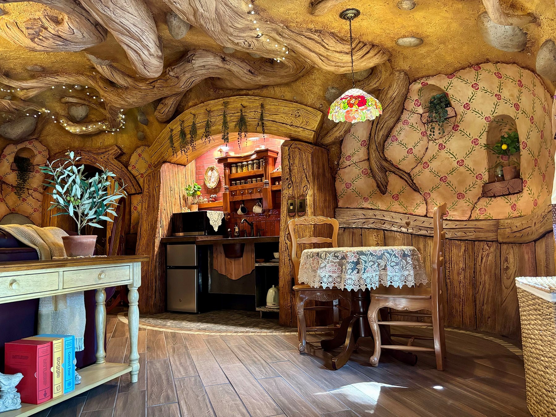 Interior of a hobbit-style room with carved wood-grain walls, floral wallpaper, dining table with lace cloth, kitchenette, and Tiffany pendant lamp