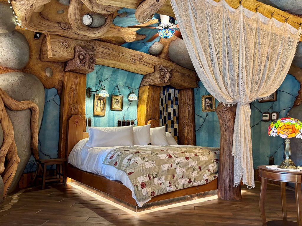 Hobbit-style bedroom with a canopy bed, floral quilt, carved log beam ceiling, blue stone walls, and stained glass lamp