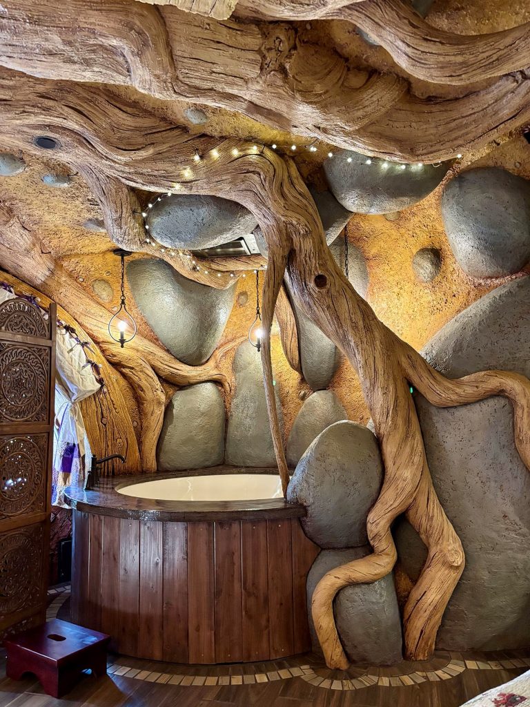 Fantasy-themed soaking tub alcove with sculpted tree root and boulder walls, string lights, and hanging candle pendants