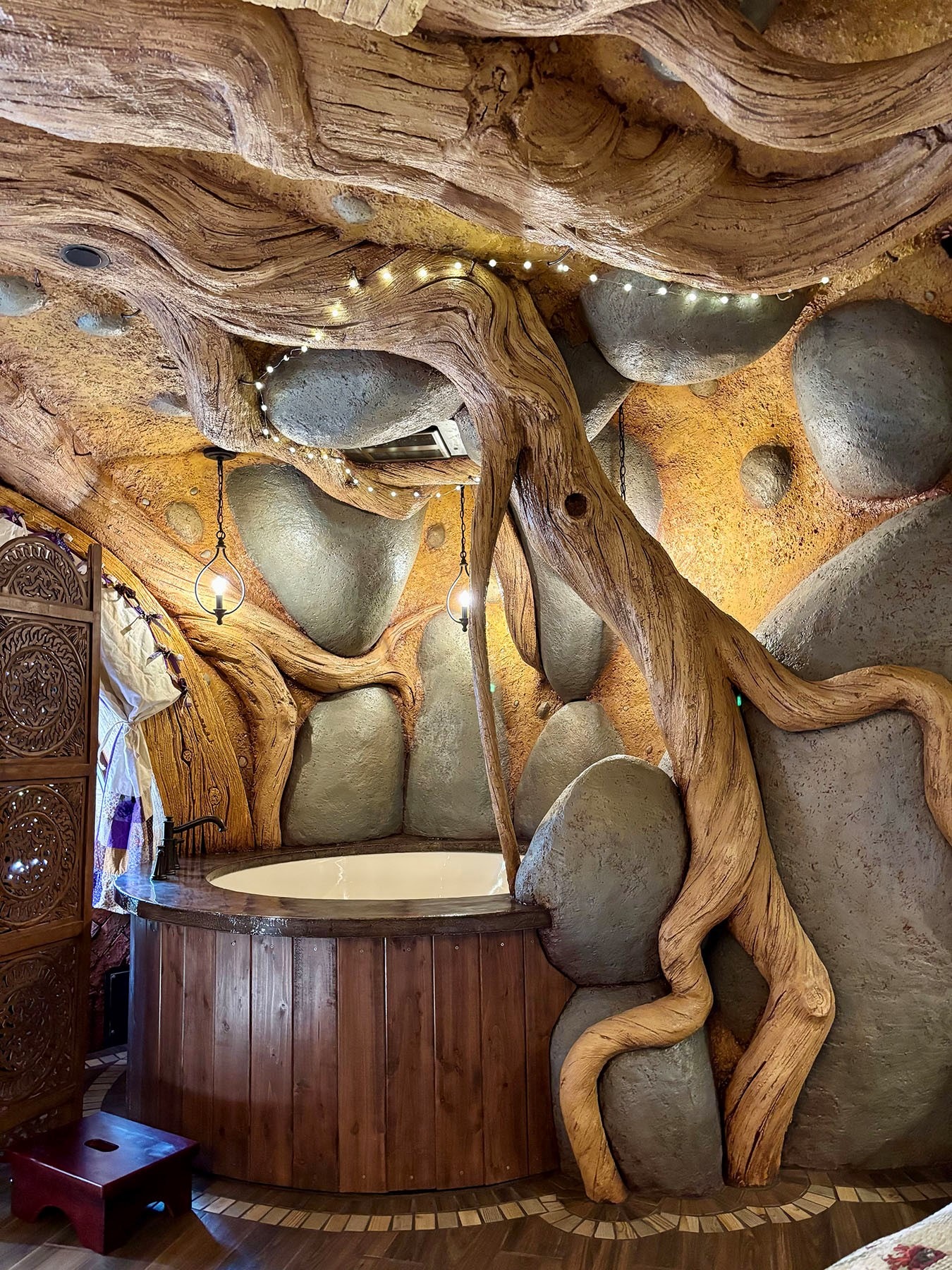 Fantasy-themed soaking tub alcove with sculpted tree root and boulder walls, string lights, and hanging candle pendants