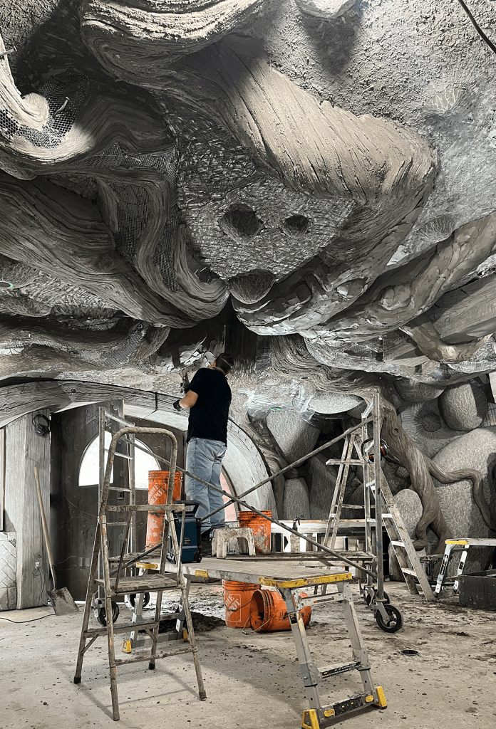 Person hand-sculpting a textured tree root ceiling inside a fantasy-themed building under construction, surrounded by scaffolding and buckets
