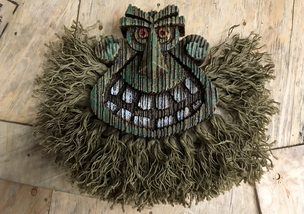 Carved wood troll face mask with red eyes and shaggy rope hair, laid flat on a wooden surface