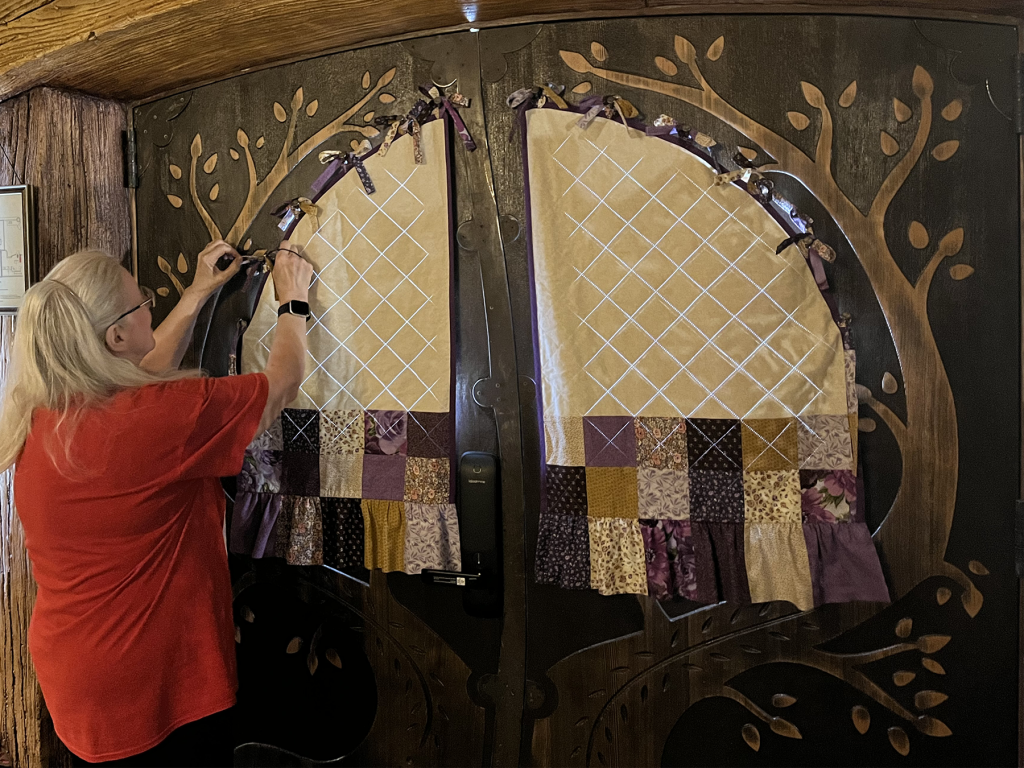 Person attaching patchwork fabric curtains to a round hobbit-style double door carved with a tree design