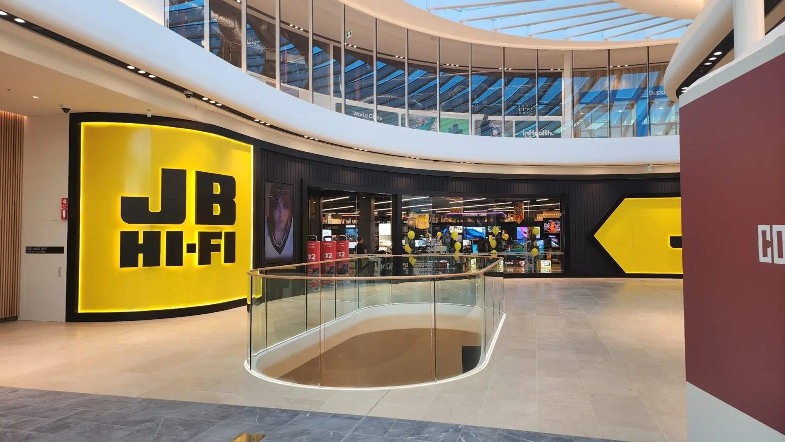 Modern JB Hi-Fi store entrance inside a shopping mall with glass railing and skylight ceiling.