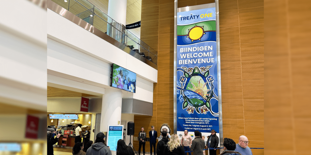 Winnipeg Airports Authority and Treaty One Nations representatives gather at the airport near the tall vertical sign. 