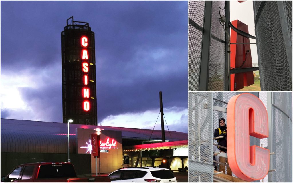 Night view of a casino with a tall vertical red "CASINO" sign and workers installing a large red letter "C" sign.
