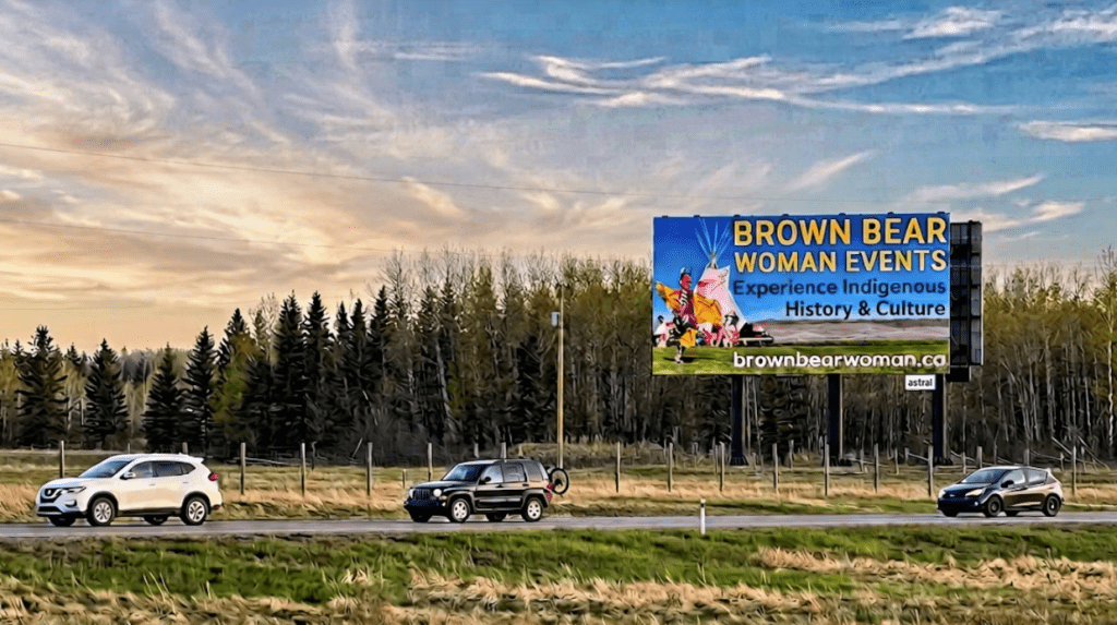 Highway scene with three cars passing by a billboard promoting Brown Bear Woman Events showcasing Indigenous history and culture.