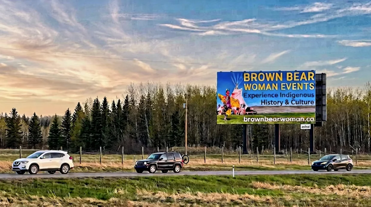Highway scene with three cars passing by a billboard promoting Brown Bear Woman Events showcasing Indigenous history and culture.