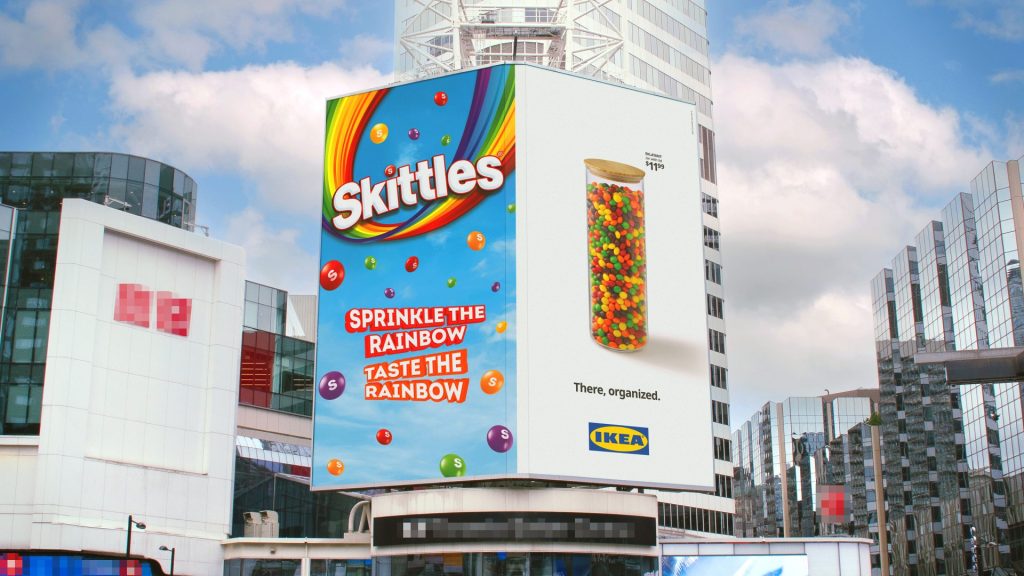 Large city billboard featuring colourful Skittles and IKEA advertisements on a modern skyscraper surrounded by reflective glass buildings under a cloudy blue sky.