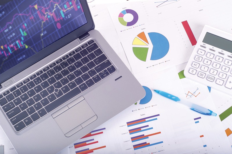 A laptop, calculator, pen, and printed charts and graphs spread across a desk.