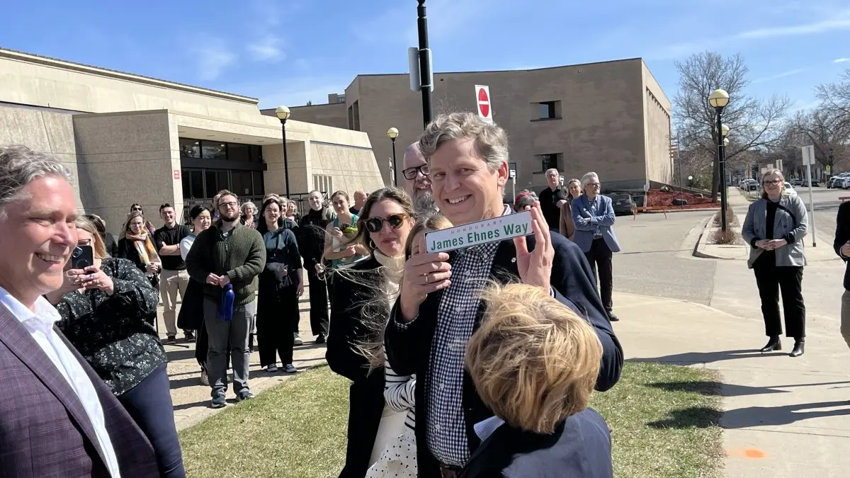 A group of people gathered outdoors, with James Ehnes holding a "James Ennes Way" street sign on a sunny day.