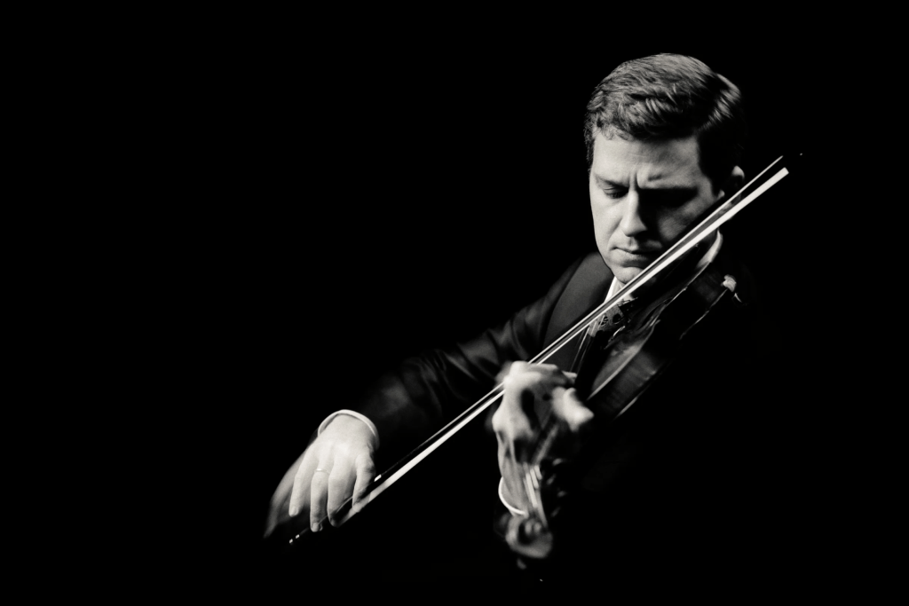 Black and white image of James Ehnes in a suit playing the violin against a dark background.