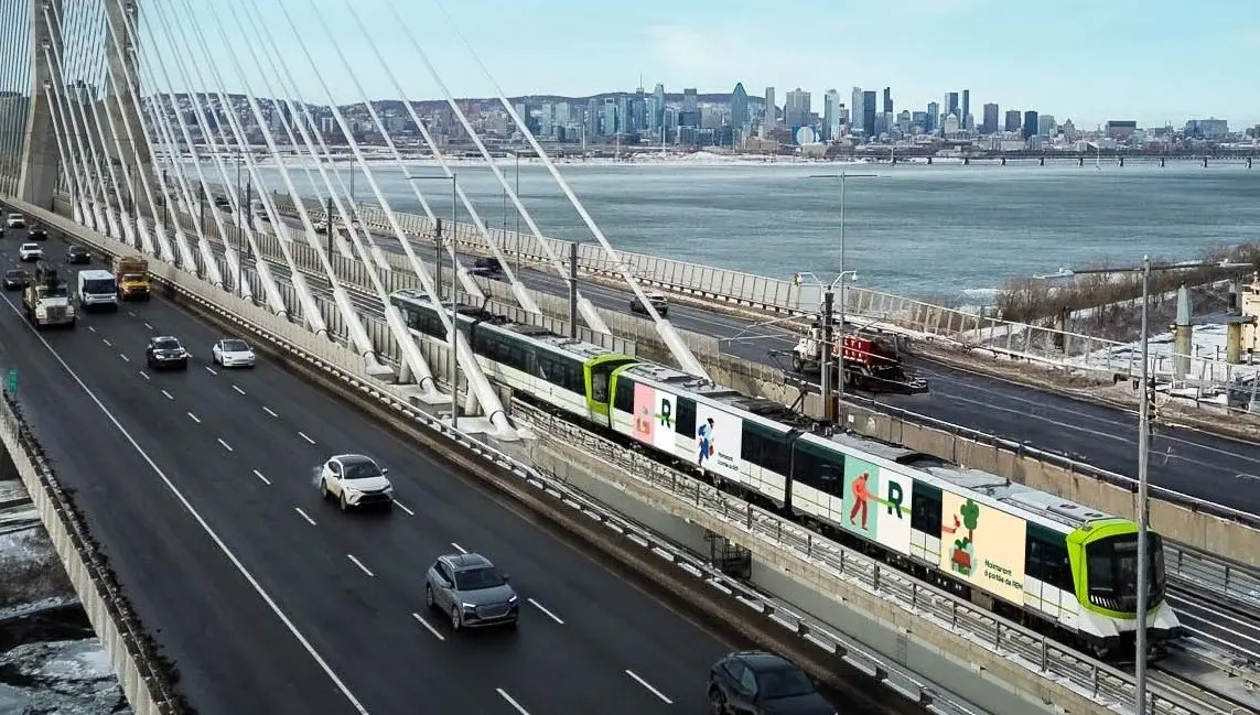 Train traveling on a cable-stayed bridge over a river, with cars on an adjacent highway and a city skyline in the background.