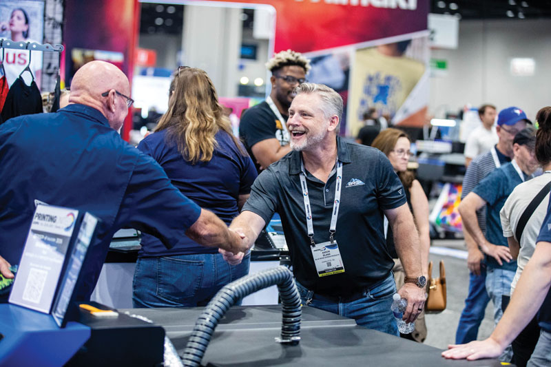 Two men shake hands and smile at a trade show booth on the PRINTING United Expo show floor.