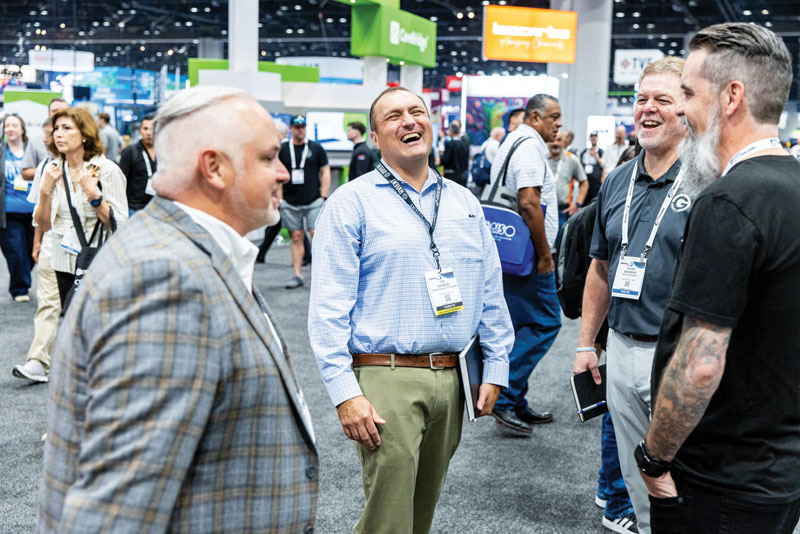 A group of men in lanyards laugh and talk together on the PRINTING United Expo show floor.