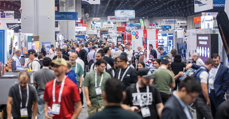 A wide view of a crowded convention hall aisle lined with exhibitor booths and signage at PRINTING United Expo.