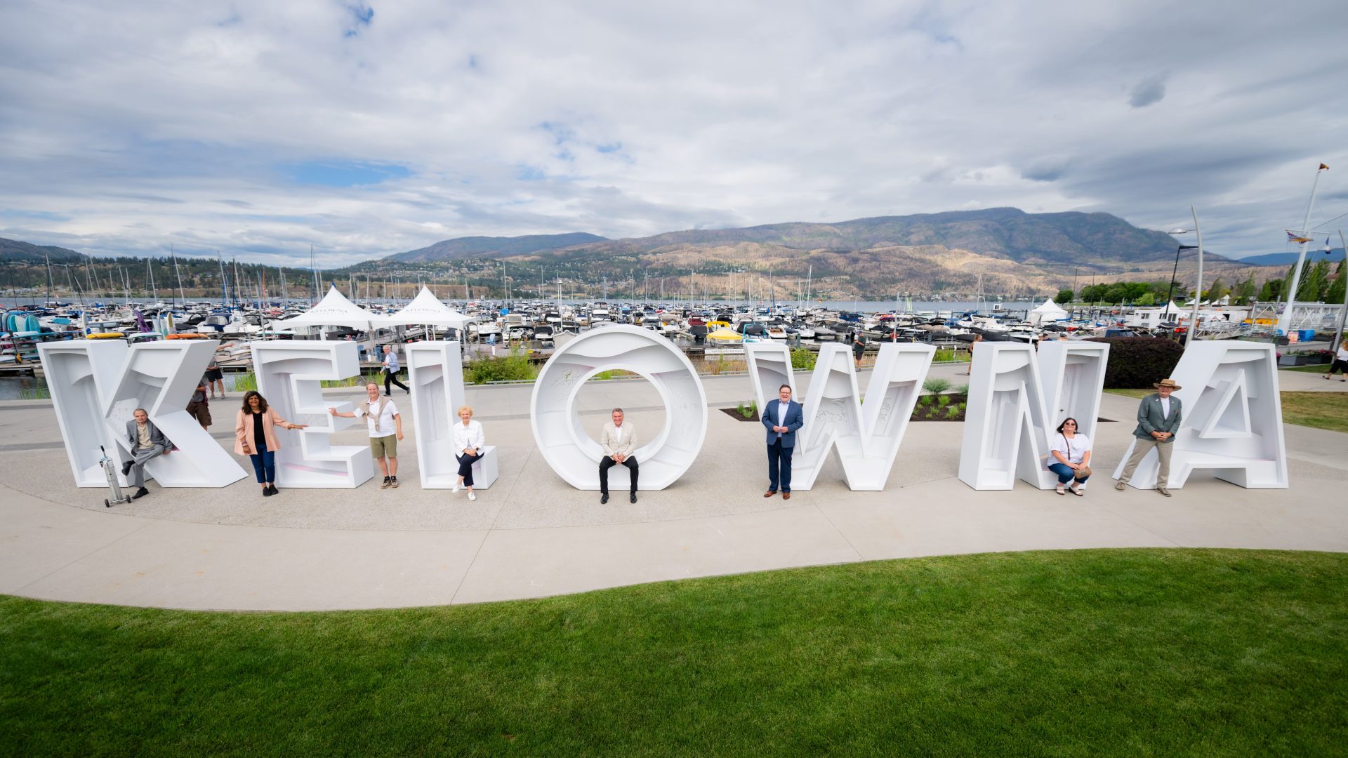 The mayor and city council pose in front of and inside the giant white "KELOWNA" letter sign on the waterfront, with a boat-filled marina and mountains behind them.
