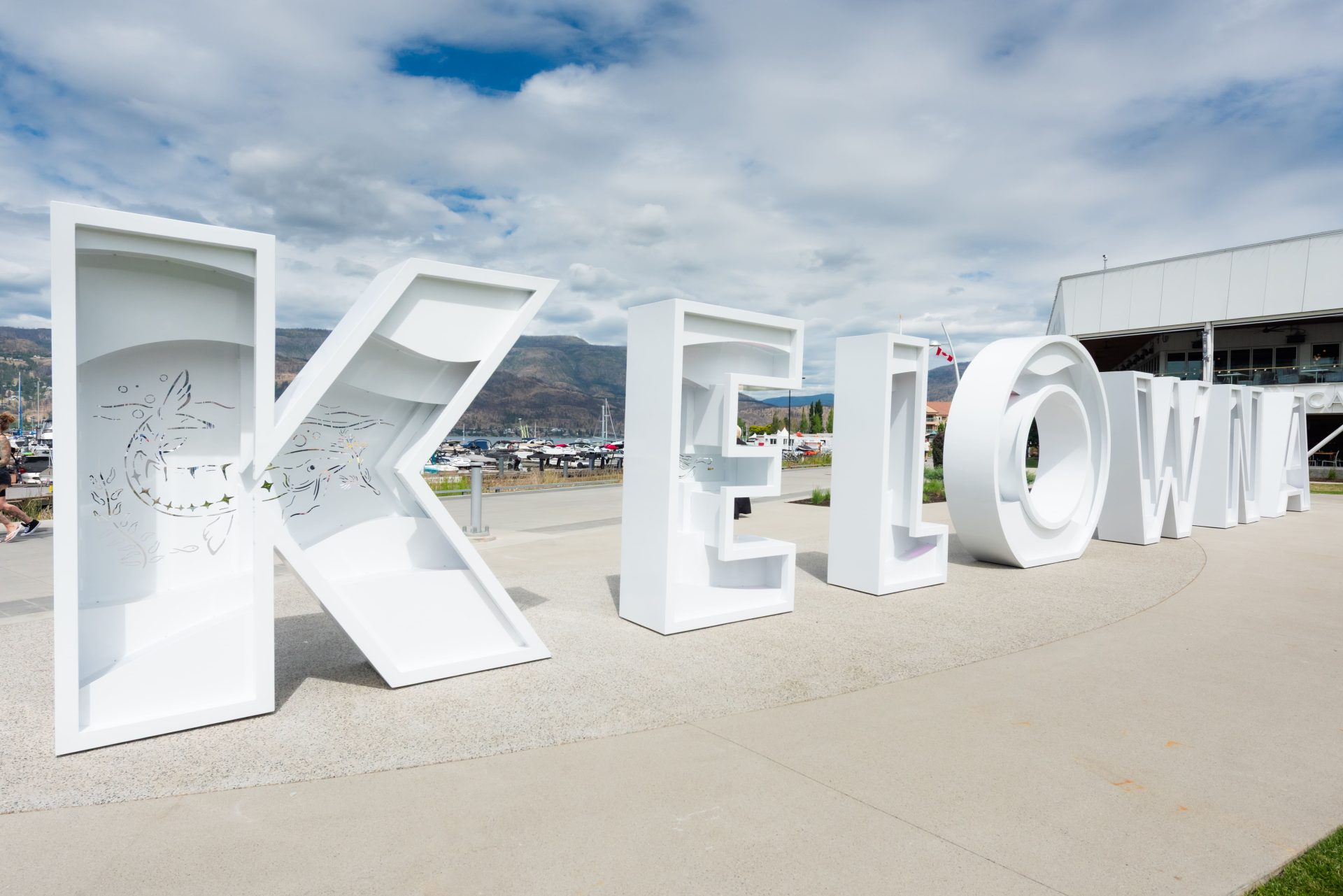 A close-up view of the large white "KELOWNA" letter sculpture along the waterfront, showing the "K" and "E" with laser-cut nature designs, set against a marina full of boats and mountains in the background.
