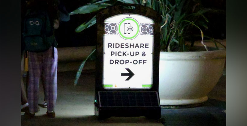 An illuminated solar-powered A-frame sign reading "RIDESHARE PICK-UP & DROP-OFF" with a directional arrow, set up outdoors at night near a planter.