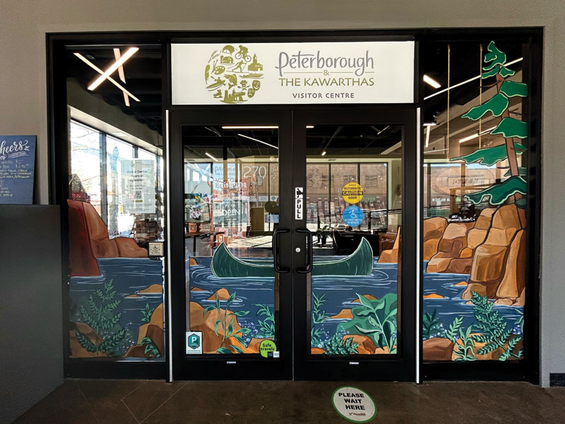 Storefront windows of the Peterborough & The Kawarthas Visitor Centre feature a painted canoe, pine tree, and rocky shoreline mural.