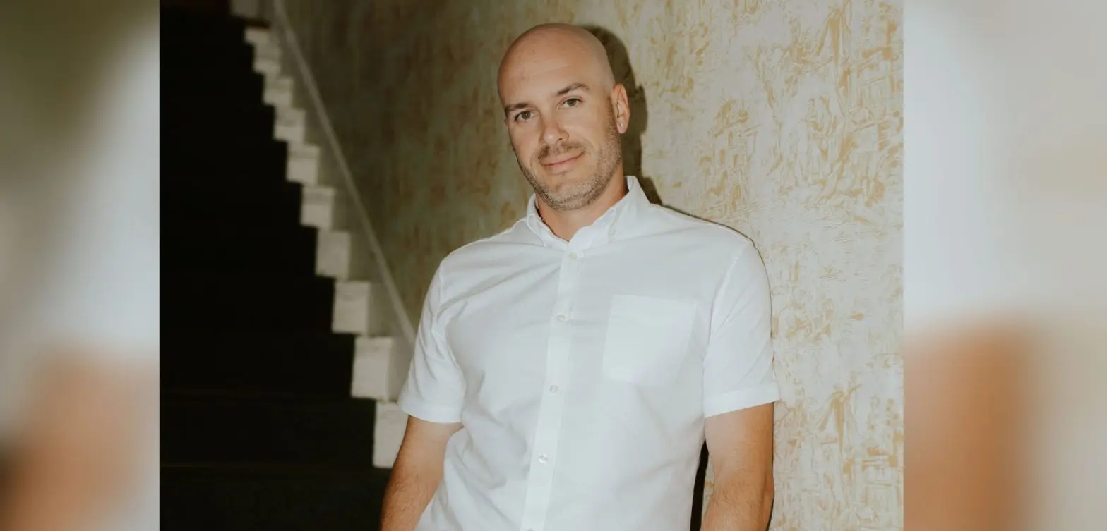 Portrait of Kort Mehrle in a white shirt, leaning against a wall with patterned wallpaper.