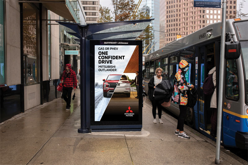Digital bus shelter ad for Mitsubishi Outlander PHEV reading 'Gas or PHEV: One Confident Drive,' next to a city bus at a downtown stop.