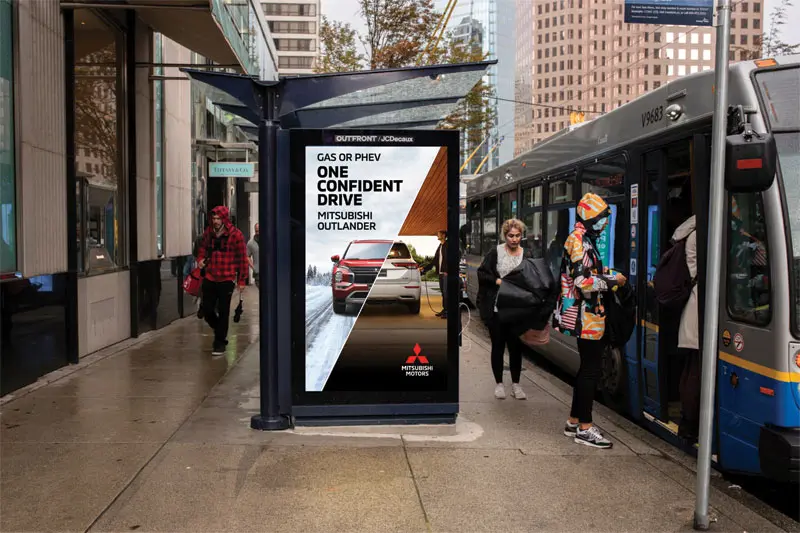 Digital bus shelter ad for Mitsubishi Outlander PHEV reading 'Gas or PHEV: One Confident Drive,' next to a city bus at a downtown stop.