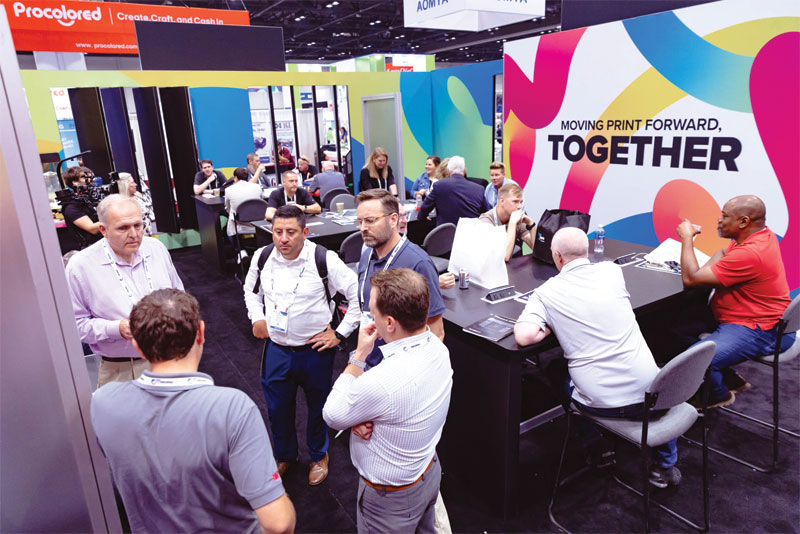 Attendees gather at tables and booths near a large "Moving Print Forward, Together" sign at the expo.