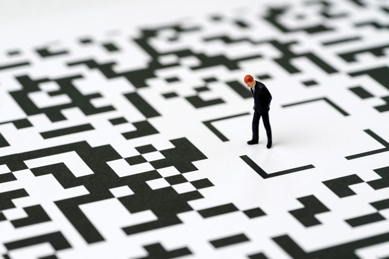 A miniature figurine of a businessman stands and looks down while walking through an oversized QR code pattern, like a maze.