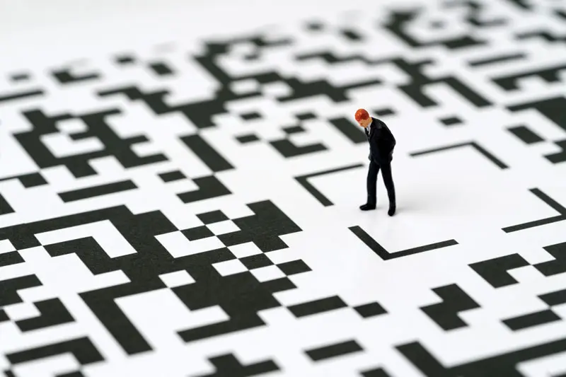 A miniature figurine of a businessman stands and looks down while walking through an oversized QR code pattern, like a maze.