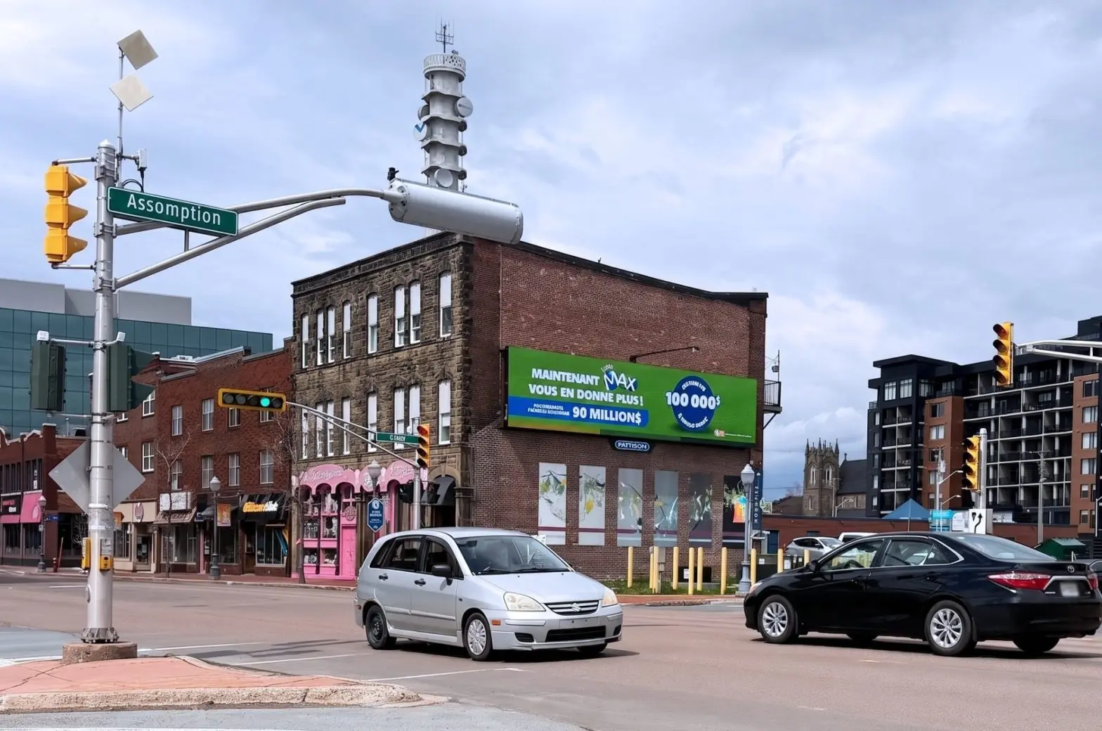 A green Pattison digital billboard advertising Loto-Québec’s “Max” jackpot, mounted on a brick building at the corner of Assomption and Riverside, with traffic signals and city traffic in the foreground.