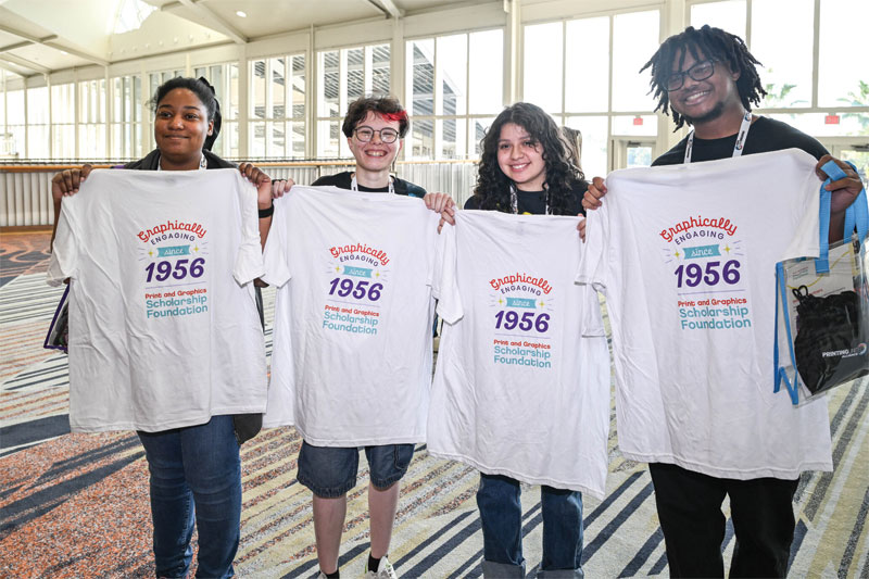 Four smiling students hold up matching white t-shirts reading "Graphically Engaging Since 1956, Print and Graphics Scholarship Foundation."
