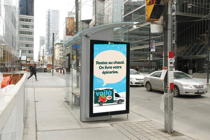 Bus shelter ad in French for Voilà by IGA grocery delivery, reading 'Restez au chaud. On livre vos épiceries,' with a delivery van illustration, on a city street.