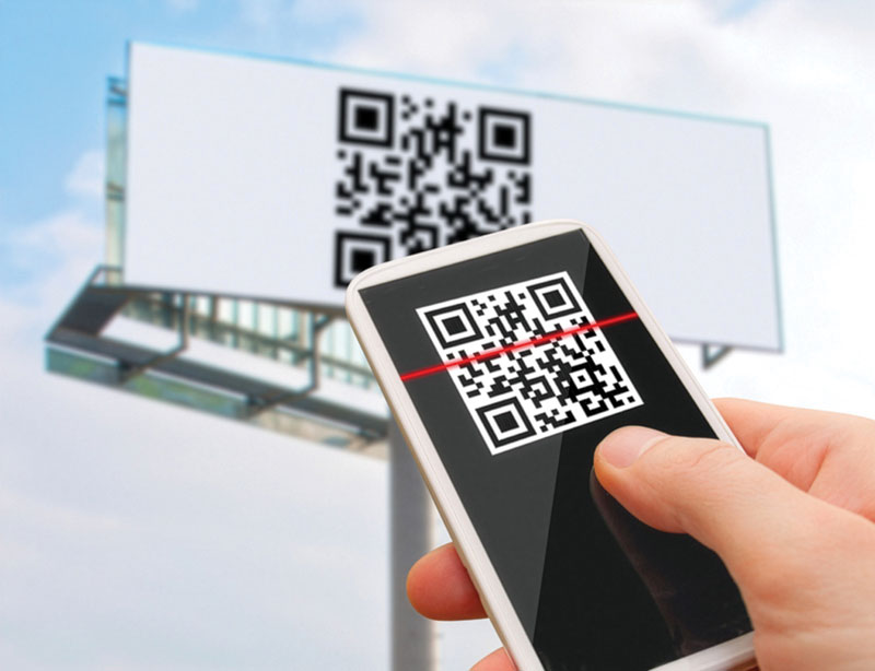 A hand holds a smartphone scanning a QR code displayed on a large outdoor billboard against a blue sky.