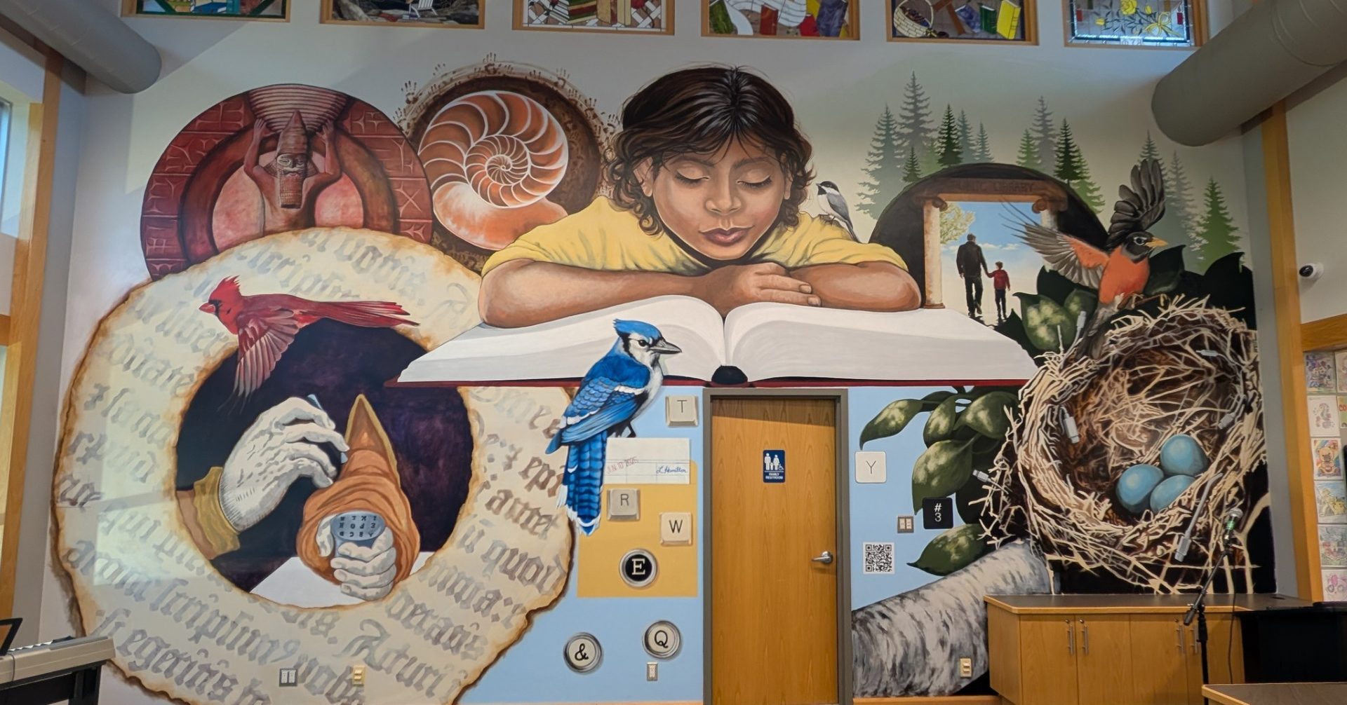 Library mural of a child reading a giant open book, surrounded by birds (blue jay, cardinal, chickadee, robin with nest), pine trees, seashells, and old library-card imagery, painted around a restroom door.