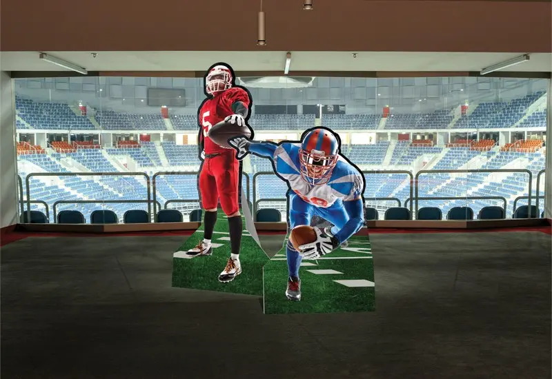Life-size cardboard cutouts of two football players printed on grass-textured bases, displayed in a stadium suite.