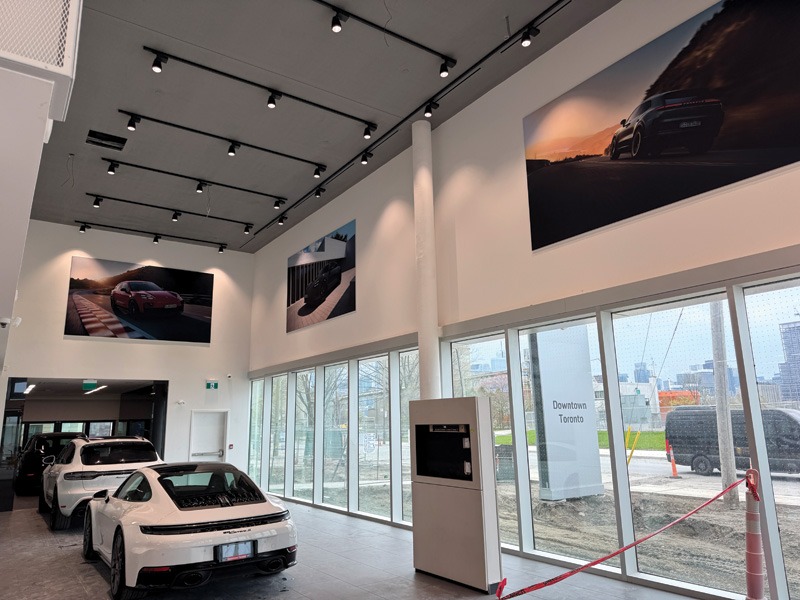 Porsche showroom interior with large wall-mounted lifestyle photo panels above two parked cars and floor-to-ceiling windows.