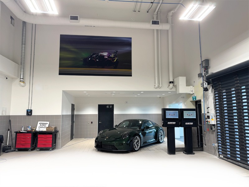 Porsche service bay with a large racing-image wall panel above a dark green 911 GT3 on an alignment lift.