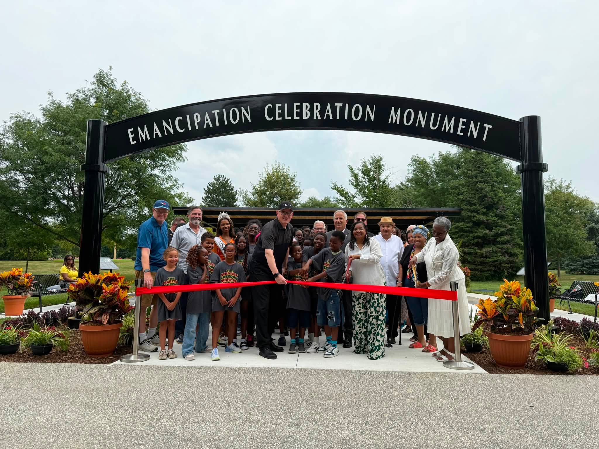 Ribbon-cutting ceremony under an arched sign reading "Emancipation Celebration Monument," with a crowd of officials, community members, and children.