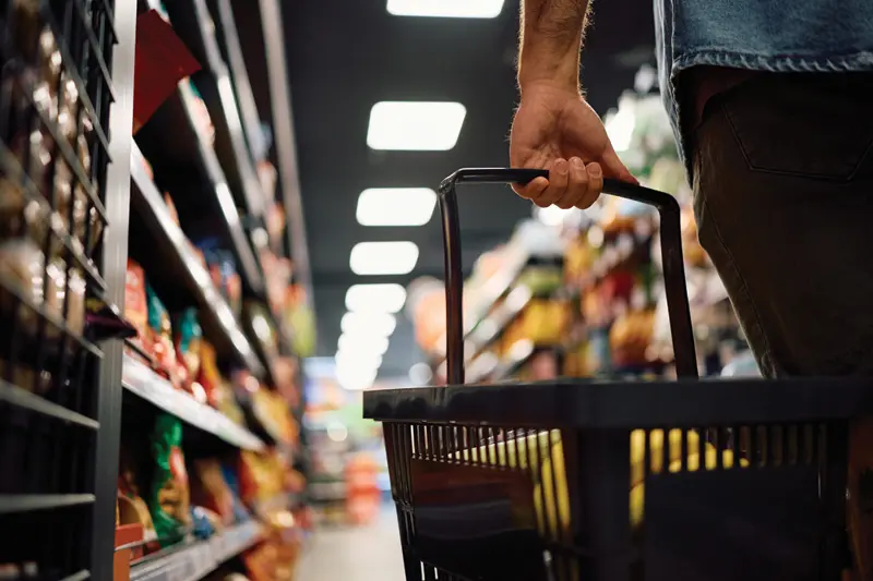 Close-up of a shopper's hand on a basket handle walking down a stocked store aisle.